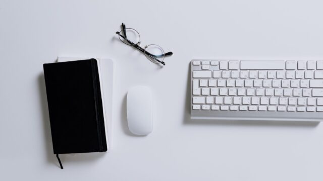 Białe biurko z klawiaturą, myszką, okularami i czarnym notesem.