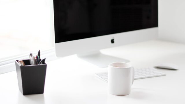 Białe biurko z komputerem iMac, klawiaturą i białym kubkiem.