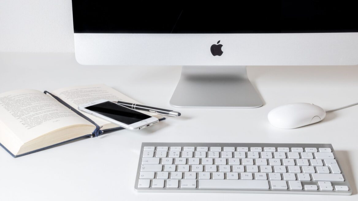 Komputer iMac, klawiatura, mysz, iPhone i książka na białym biurku.